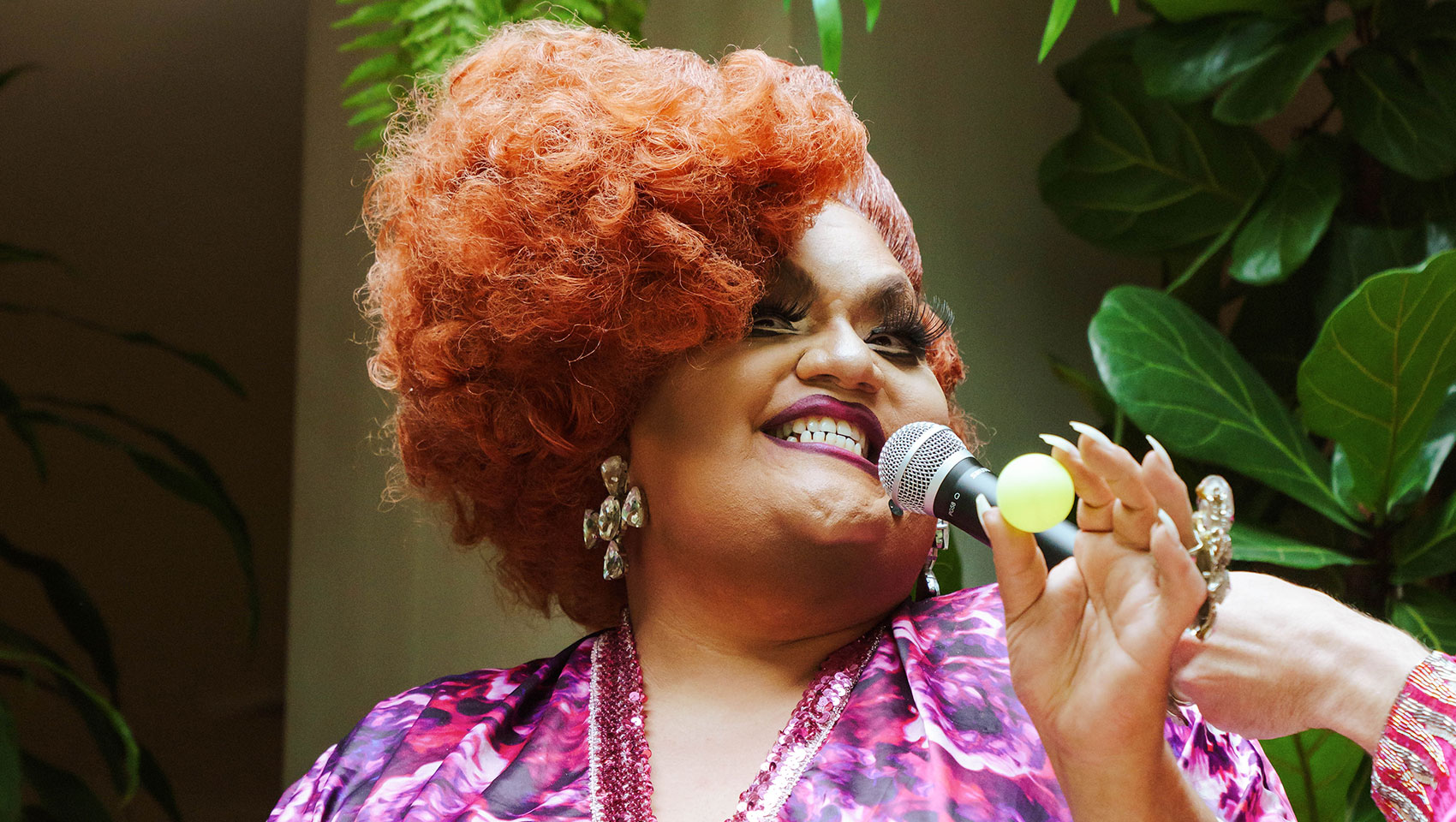 Drag queen with red wig and purple floral mumu with microphone